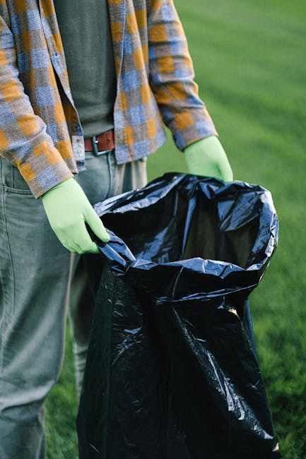 A person wearing a green long-sleeve shirt, a plaid jacket with yellow, blue, and gray patterns, and matching pants, is holding open a large black rubbish bag lined with a shiny interior. The individual is wearing light green gloves and standing outdoors on a grassy area, with a blurred background of green grass or a park-like setting. The focus is on the person's hands and the rubbish bag, which appears to be prepared for waste collection or disposal, reflecting typical practices in private rubbish removal services such as those provided by Rubbish Removal Maida Vale. The scene suggests an on-site rubbish collection activity, emphasizing cleanliness and waste management in an outdoor environment, relevant to independent waste handling and rubbish removal efforts.