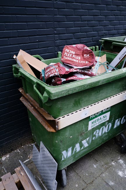 A green wheeled dumpster filled with multiple red and black bags of garden waste, including soil and compost bags labeled 'FLEXI PLUS.' The dumpster is positioned on a paved surface against a dark blue brick wall, with part of a white waste container visible below. Nearby, a small metal grate is leaning against the dumpster, and the scene appears to be outdoors, possibly in a residential or commercial garden or back alley. The image depicts private waste collection, consistent with independent rubbish removal services like those provided by Rubbish Removal Maida Vale, and emphasizes on-site clearance of garden refuse.