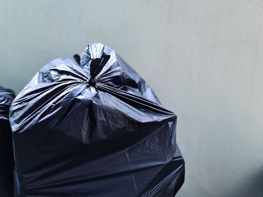 Close-up of a tightly tied black plastic rubbish bag with a glossy, slightly wrinkled surface, placed against a plain light-colored wall. The bag appears full and is situated in an indoor or enclosed environment, possibly awaiting collection for waste disposal or recycling. The background is neutral and unobtrusive, focusing attention on the bag itself, which is associated with private waste handling services such as those provided by Rubbish Removal Maida Vale. The scene emphasizes the typical appearance of rubbish prepared for collection, highlighting the practicality of using sealed plastic bags for rubbish removal tasks. The lighting is even, with minimal shadows, ensuring the details of the bag’s texture and creases are clear, supporting an accurate, accessible description suitable for individuals relying on image descriptions related to rubbish removal services.