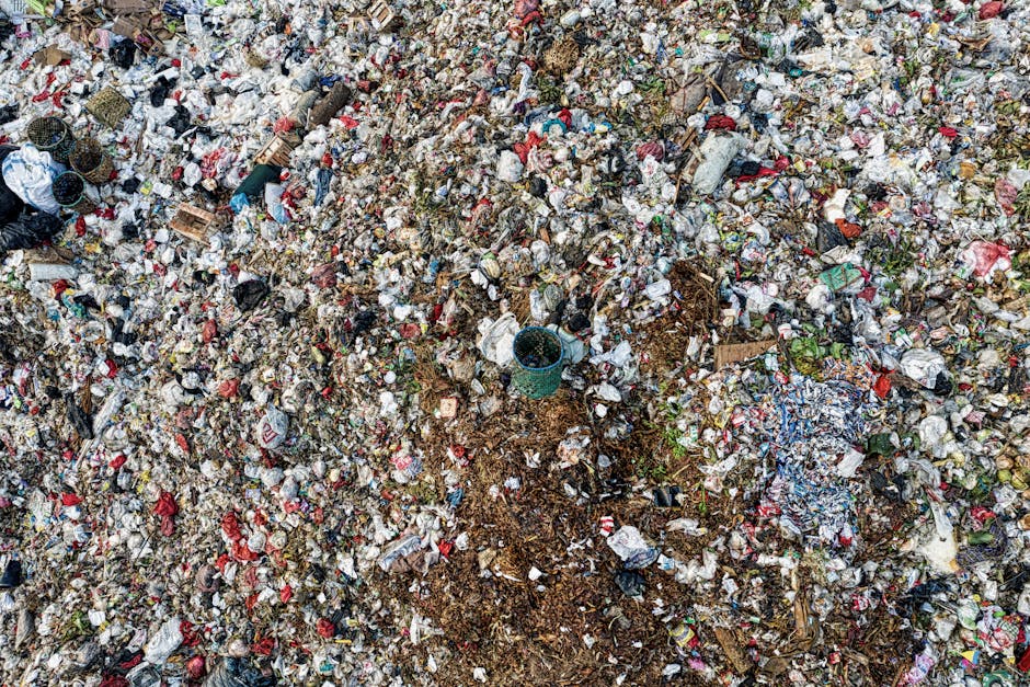 A green wheeled dumpster filled with multiple red and black bags of garden waste, including soil and compost bags labeled 'FLEXI PLUS.' The dumpster is positioned on a paved surface against a dark blue brick wall, with part of a white waste container visible below. Nearby, a small metal grate is leaning against the dumpster, and the scene appears to be outdoors, possibly in a residential or commercial garden or back alley. The image depicts private waste collection, consistent with independent rubbish removal services like those provided by Rubbish Removal Maida Vale, and emphasizes on-site clearance of garden refuse.
