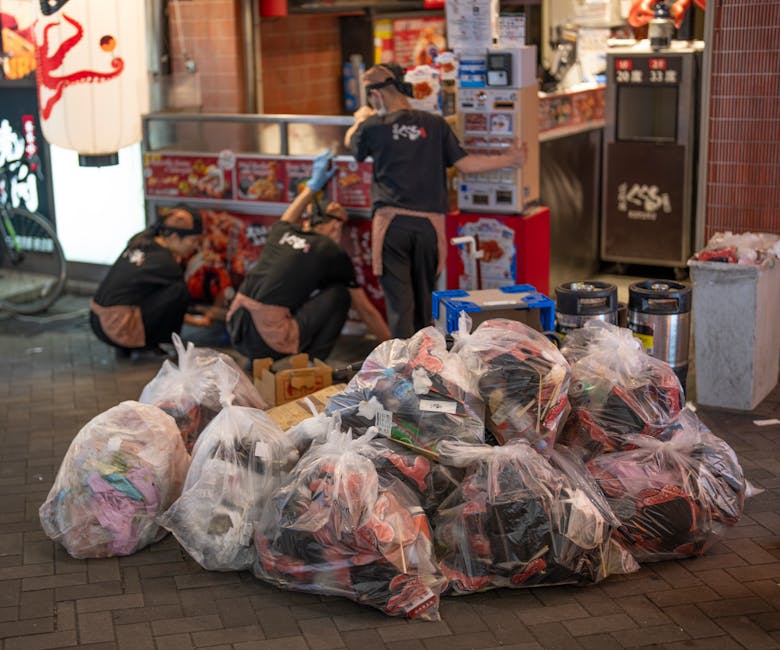 A collection of overflowing rubbish bins and scattered waste bags on a paved sidewalk in front of a commercial building and parking area. The scene includes a large grey recycling container marked for mixed paper and card, with its lid open, revealing various crumpled papers, cardboard, and general waste. To the right are black and red bins, also filled and with waste spilling out, including black plastic bags and flattened cardboard boxes. Surrounding the bins on the ground are loose cardboard boxes, paper packaging, plastic bags, and miscellaneous trash, some partially collapsed or torn. Behind the waste area, a metal railing runs along the edge of the sidewalk, and a parked car is visible on the left. The building in the background has a blue metal staircase and signage, with storefronts beneath. The overall scene depicts a state of unmanaged refuse, indicative of a private waste collection or an area awaiting rubbish removal, with environmental concern over cluttered and overflowing waste receptacles.