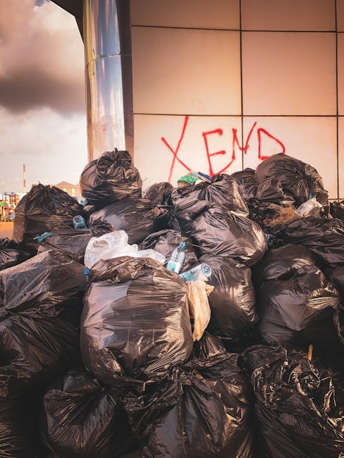A large pile of black plastic rubbish bags is stacked against a tiled wall exterior of a building. The bags are crumpled and vary in size, with some appearing full and tightly tied, while others are slightly looser. Among the black bags, a few transparent plastic bottles and small pieces of discarded packaging are visible, suggesting mixed household waste. The wall behind the rubbish piles features beige tiles with a reddish graffiti tag reading 'XEND' painted on it. In the background to the left, part of a cloudy sky is visible, indicating an outdoor setting during daytime. The scene appears to be an instance of overfilled private rubbish collection or waste clearance, likely handled by a specialist service such as Rubbish Removal Maida Vale, supporting on-site or independent disposal without authority collection. Overall, the image depicts a typical rubbish accumulation spot requiring professional waste management to facilitate proper clearance and disposal.