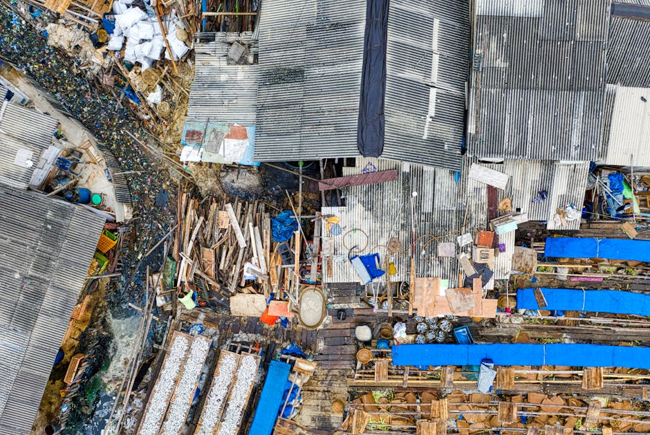 An aerial view of a cluttered outdoor area showing a variety of discarded materials and waste. In the foreground, there are wooden planks, pallets, and broken furniture arranged haphazardly with some pieces leaning against makeshift structures. Adjacent to this, a patch of uneven ground contains scattered debris, including plastic containers, metal objects, and bits of fabric. To the right, there are several long blue tarpaulins or covers stretched over wooden frameworks, possibly used to shelter stored items or waste. The background features a series of corrugated metal roofs, some partially rusted, covering sheds or storage units. The scene is illuminated by natural light, emphasizing the rough textures of the wood, metal, and plastic, and creates a sense of disorganized accumulation typical of private waste handling or site clearance scenarios. This image exemplifies the type of waste often dealt with during rubbish removal services, highlighting the need for professional clearance to manage such clutter efficiently. Rubbish Removal Maida Vale regularly handles similar outdoor rubbish and waste clearance tasks to facilitate clean and safe environments.