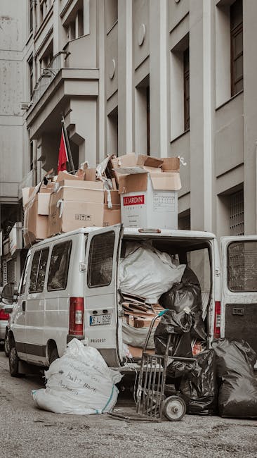 A narrow, urban alleyway filled with accumulated rubbish and debris, with a large black garbage bag prominently in the foreground amid a scattered collection of discarded items. Behind the bag, pallets and piles of cardboard boxes, paper, and other waste materials are stacked haphazardly against a backdrop of weathered brick walls and graffiti-covered surfaces. The alley is bordered on the right by a grey building with metal security bars over window openings, while on the left, a rusty, dark-toned structure leans into the space. A small trash bin and miscellaneous refuse lie on the ground to the left, with some debris strewn across the uneven, stained pavement. Bare tree branches extend over the alley, hinting at an outdoor setting possibly nearby residential or commercial buildings. The scene appears to be a collection point for waste awaiting removal, illustrating the need for proper rubbish clearance, which companies like Rubbish Removal Maida Vale offer as an alternative to public or municipal waste disposal methods.