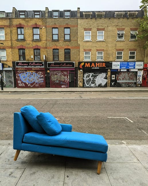 A bright blue fabric-upholstered chaise lounge with a slightly curved seat and matching cushions positioned on a concrete sidewalk in front of a row of commercial buildings. The chaise features smooth, vibrant upholstery with visible stitching, supported by four wooden tapered legs. The scene is outdoors, with a street behind the furniture showing faint markings on the tarmac. The background comprises a multi-story brick building with several windows, some with white blinds, and storefronts with metal roller shutters, many of which are covered in graffiti and tags. Overhead, a cloudy sky casts diffuse natural light on the scene. The overall setting suggests an urban environment where independent collection or private waste handling, such as furniture removal by Rubbish Removal Maida Vale, might be involved in the context of urban rubbish clearance.
