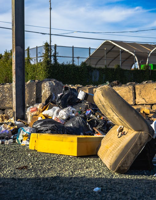 A pile of mixed waste including black plastic bin bags, cardboard boxes, and discarded furniture items such as an old, worn out car tire and a yellow plastic container, all scattered on the ground of an outdoor area. The waste is placed directly on a gravel surface beside a low stone wall and nearby a metal pole with wires overhead. In the background, there are trees, a hedge, and structures that include a partially visible curved metal or plastic shelter, suggesting a residential or semi-industrial environment. The scene appears to be during daylight with clear blue skies, highlighting the need for proper rubbish removal and potentially supporting services like private waste collection that aim to prevent such unmanaged accumulation that can lead to delays or issues with local rubbish disposal routines. Rubbish Removal Maida Vale operates in environments involving on-site clearance and independent waste handling, as reflected here with visible unmanaged waste that could require professional removal services for effective and timely cleanup.