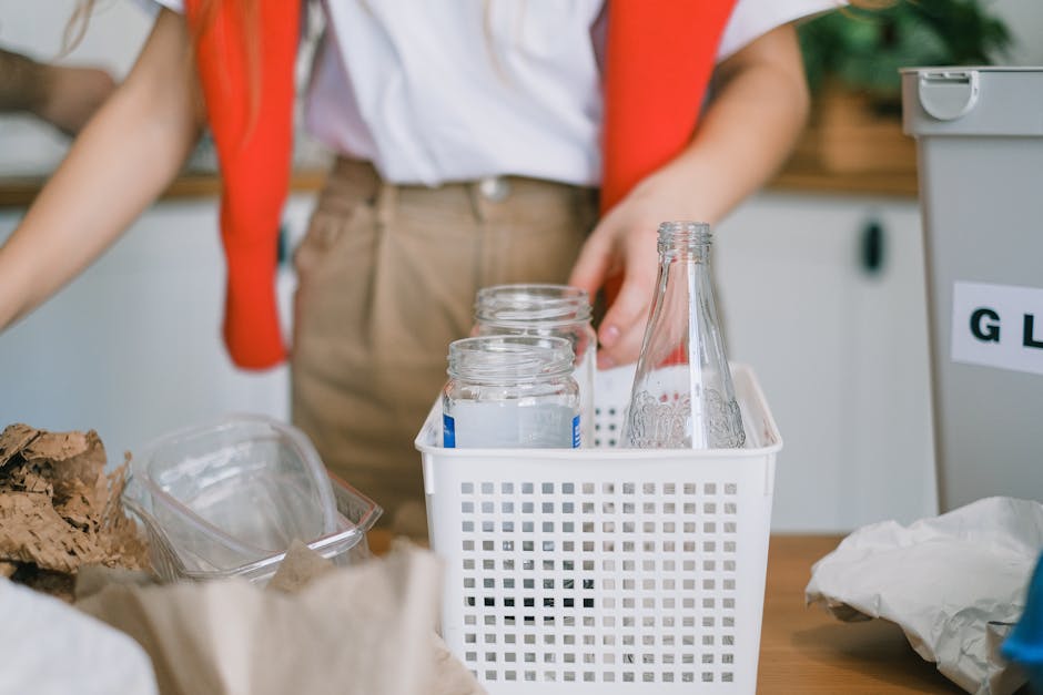 A person wearing a white shirt and beige trousers is standing at a cluttered table in a recycling or waste collection area. Their right arm is slightly bent, holding a white plastic basket containing four empty glass bottles, which appear to be soda or beer bottles, with a transparent, smooth surface and metal screw caps removed. The bottles are clear and free of labels, positioned upright in the basket, while an additional glass bottle, larger with a longer neck, is leaning at an angle beside them. The table surface has various discarded items, including crumpled brown paper, a transparent plastic container, and other recyclable or waste materials. In the background, there are blurred elements of a grey storage bin with a label partly visible, and a green leafy plant or foliage can be seen, indicating an indoor or semi-outdoor environment dedicated to waste sorting or re-use activities. The scene demonstrates a focus on the handling of glass recyclables, reflecting an aspect of private waste management or alternative rubbish removal options conducted by Rubbish Removal Maida Vale.