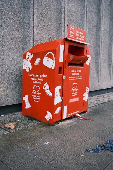 A collection of flattened and crumpled cardboard boxes and paper packaging materials are stacked in front of a metal shipping container, which has a blue sign reading 'RECYCLING ONLY' with a green recycling symbol. The boxes vary in size, with some showing printed branding, and are a mix of brown kraft and recycled paper textures, with some appearing torn or bent. The cardboard packaging sits on top of or leaning against brown and blue recycling bins designated for paper waste, which are positioned on a paved surface. Above the containers, part of a metallic framework or shelving is visible, suggesting an organized commercial or industrial waste collection area dedicated to recyclable materials. Natural daylight illuminates the scene, highlighting the corrugated surface of the containers and the torn edges of the cardboard, emphasizing the focus on waste sorting and disposal for recycling purposes. Rubbish Removal Maida Vale specializes in private waste handling, including the collection of recyclable packaging materials in accordance with local disposal guidelines and environmentally responsible practices.