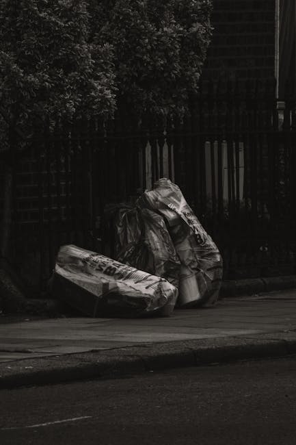 Two large black garbage bags filled with waste material are placed on a paved sidewalk beside a black metal fence, with a dense leafy shrub or tree behind the fence in the background. The bags appear crumpled and are made of plastic, with some visible printed text on the surfaces. The scene is captured outdoors in natural daylight, with minimal surrounding environment visible apart from the curb and part of a street in the foreground. The bags are positioned on the roadside, likely awaiting collection, highlighting a typical scene of private waste disposal managed by a rubbish removal service such as Rubbish Removal Maida Vale, which specialises in alternative waste handling solutions like on-site clearance and independent collection amidst urban residential environments.
