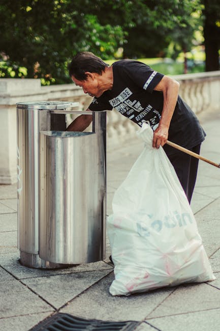An elderly man with dark hair, wearing a dark t-shirt with white text and dark trousers, is seen bending over a stainless steel public trash bin on a paved sidewalk. He is placing a white plastic bag filled with rubbish into the bin, holding a wooden stick or cleaning tool in his right hand. The background features a low stone wall, lush green trees, and a park-like environment, indicating an outdoor setting. The scene is well-lit by natural daylight, with the greenery providing a contrasting backdrop to the metallic surface of the waste receptacle. This image exemplifies responsible waste disposal and may reflect private or independent rubbish removal activities, often associated with community cleaning efforts or individual waste management in urban outdoor spaces, aligning with the services of Rubbish Removal Maida Vale.