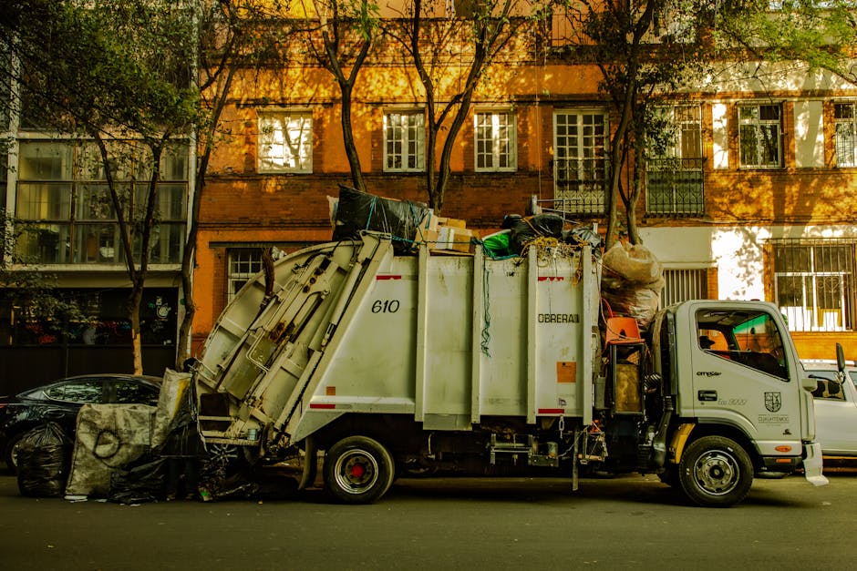A side view of a commercial waste collection vehicle parked on a city street in front of a multi-storey residential building with orange brick walls and white-framed windows. The vehicle, marked with the number 610, has an open top loaded with various types of rubbish, including large black garbage bags, flattened cardboard boxes, plastic containers, and loose debris, indicating it is engaged in on-site clearance or alternative waste handling. The truck is equipped with a hydraulic system at the rear for compacting and lifting waste, and the cab is painted white with a grey logo on the door. Surrounding the vehicle, there are parked cars—one black vehicle on the left and a small white van on the right—and a row of tall, leafy trees casting shadows on the building facade and the street. The scene is illuminated by natural daylight, emphasizing the textures of the waste materials and the urban environment, with a subtle connection to rubbish removal services offered by companies such as Rubbish Removal Maida Vale.