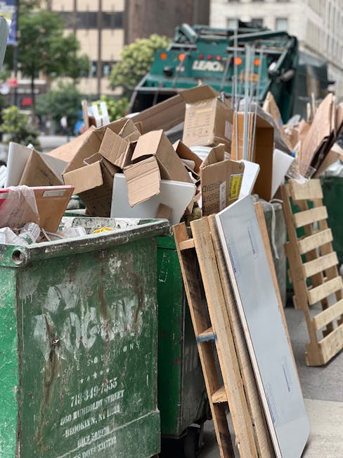 A large green waste collection bin filled with various types of construction and household debris, including multiple cardboard boxes of different sizes, some flattened and others partially assembled, along with wooden pallets leaning against the side. The cardboard is scattered and stacked atop the bin, revealing a mixture of brown and white materials. Behind the bin, a refuse collection vehicle with a teal and white colour scheme and a brand name partially visible on the side is positioned on a city street, surrounded by urban buildings, trees, and parked vehicles. The environment appears to be an outdoor urban setting during daylight hours, with natural light illuminating the scene and highlighting the textures of the cardboard, wood, and metal surfaces. This scene exemplifies a typical private waste collection scenario, where rubbish such as packaging, pallets, and miscellaneous debris is prepared for removal by a waste management service, supporting the context of independent waste handling and site clearance.