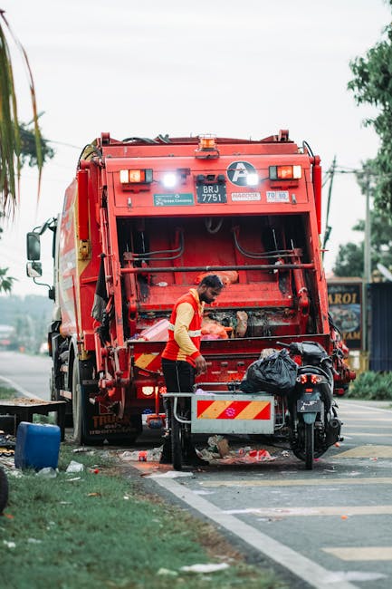 A side view of a commercial waste collection vehicle parked on a city street in front of a multi-storey residential building with orange brick walls and white-framed windows. The vehicle, marked with the number 610, has an open top loaded with various types of rubbish, including large black garbage bags, flattened cardboard boxes, plastic containers, and loose debris, indicating it is engaged in on-site clearance or alternative waste handling. The truck is equipped with a hydraulic system at the rear for compacting and lifting waste, and the cab is painted white with a grey logo on the door. Surrounding the vehicle, there are parked cars—one black vehicle on the left and a small white van on the right—and a row of tall, leafy trees casting shadows on the building facade and the street. The scene is illuminated by natural daylight, emphasizing the textures of the waste materials and the urban environment, with a subtle connection to rubbish removal services offered by companies such as Rubbish Removal Maida Vale.
