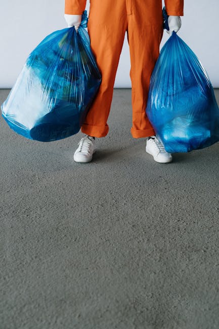 A person wearing orange work trousers and white sneakers stands on a grey surface, holding a large blue plastic rubbish bag in each hand. The bags appear full and are filled with waste materials, with the semi-transparent plastic revealing some of the contents inside. The background is a plain, neutral grey wall, emphasizing the focus on the individual and the bags. This scene suggests an act of rubbish collection or waste disposal, possibly related to private waste removal services offered by Rubbish Removal Maida Vale, highlighting an on-site clearance or independent rubbish collection process commonly seen in waste management practices in Maida Vale.