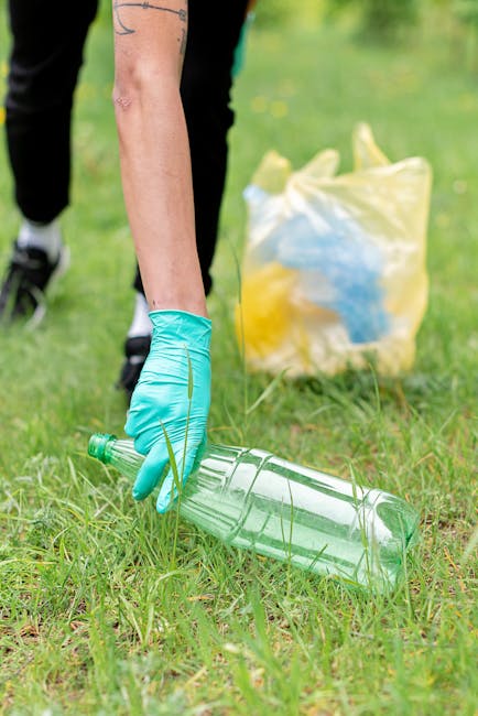 Several black plastic garbage bags are scattered on a grassy area in front of a residential building, with some bags leaning against the trunks of leafless trees. The building behind features a modern design with large windows and a combination of light grey and dark grey exterior panels. The scene is set outdoors, with overcast lighting creating a neutral, subdued atmosphere. The bags are likely filled with domestic waste, and their placement suggests an informal rubbish collection or disposal scene, which a rubbish removal service like Rubbish Removal Maida Vale might handle as part of an alternative waste removal solution. The environment indicates an urban or suburban setting, with a pavement running along the field and trees that are seasonally bare, possibly during late autumn or winter. Overall, the image captures the practical aspect of waste management outside a residential block, emphasizing the importance of organized rubbish collection to maintain cleanliness and tidy outdoor spaces.