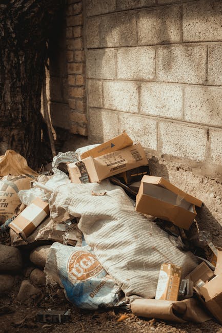A pile of mixed waste materials situated against a brick wall outdoors. The refuse includes flattened cardboard boxes with printed labels, large woven fabric or sack-like materials, and plastic bags filled with various debris. The cardboard boxes vary in size and are partially torn or open, revealing their contents. The fabric or sack material appears textured with a coarse surface and light beige color. The scene is shaded, with natural outdoor lighting. The waste is positioned on uneven ground that includes small rocks and dirt, with a large tree trunk visible to the left and a brick wall acting as a backdrop. The area suggests a temporary or informal dumping spot, which could be handled through private rubbish removal services like Rubbish Removal Maida Vale, offering an alternative to standard local authority collection for such outdoor clutter.