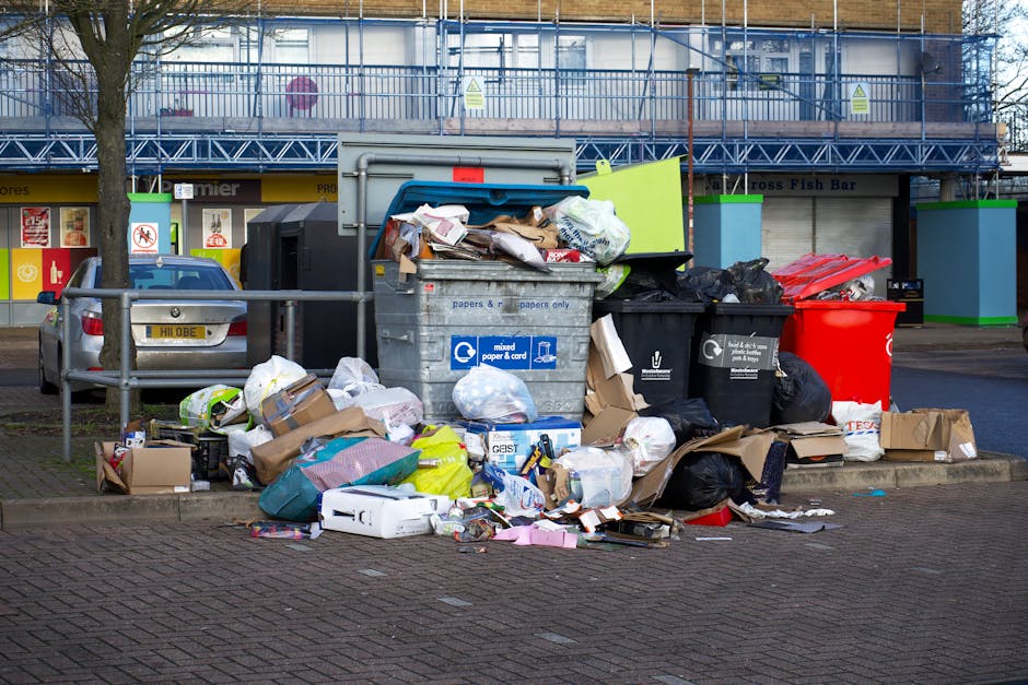 A large collection of overflowing rubbish and waste materials is piled on a paved area, including black garbage bags, cardboard boxes, paper waste, and miscellaneous packaging materials. The waste is scattered across the curb, with some items spilling onto the street, indicating an accumulation of refuse awaiting collection. Behind the waste is a row of waste bins, some made of metal, in various colours such as black, red, and black plastic, with one bin lid open and filled with discarded papers and packaging. To the left, a silver car is parked beside a metal rail, with a tree and a storefront with signage and closed shutters visible in the background. The scene suggests an urban environment, with an emphasis on the need for rubbish removal services, potentially highlighting the importance of regular unattended waste collection or private waste disposal options rather than municipal services. The setting appears to be outdoors during daylight hours, with natural light illuminating the scene clearly, emphasizing the varied textures of waste materials and the surroundings, in line with visual descriptions used in waste management contexts.