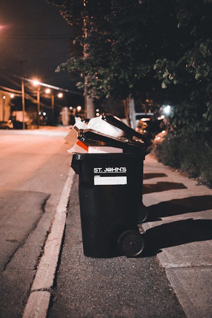 A black wheeled rubbish bin labeled 'St. John's' is positioned on a sidewalk at night, filled with a variety of discarded waste materials including brown cardboard boxes, plastic bags, and paper. The bin is situated near a curb along a quiet street illuminated by streetlights, with a row of trees and bushes visible on the right side. In the background, blurry parked cars and additional streetlights can be seen, casting a warm glow over the scene. The debris and waste inside the bin suggest it is used for on-site clearance or waste collection services, which may be an alternative to council rubbish collection, aligning with independent rubbish removal practices. The environment appears residential, with the bin placed for collection or disposal services, supporting the need for professional waste management or rubbish removal specialists like Rubbish Removal Maida Vale.